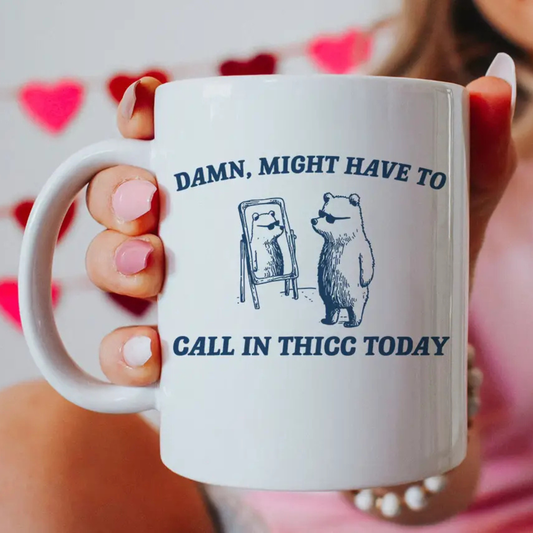 White mug with bear illustration and text held by a person with pink nail polish against a blurred background.